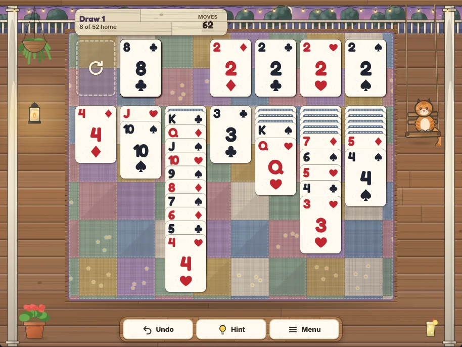 Actual Porch Swing Solitaire gameplay in a desktop browser: Draw 1 with 8 of 52 cards home after 62 moves, every Ace and 2 on the four foundations, an empty stock ready to turn the waste back over, the 8 of clubs on the waste, and seven columns on a patchwork quilt, one holding a long run from the King of clubs down to the 4 of hearts in alternating colors. Biscuit the ginger cat sits on a porch swing beside the table, with Undo, Hint and Menu on a wooden bench below.